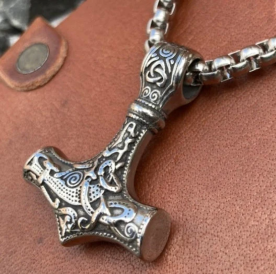 An angled view of a solid looking pendant in the shape of Thor's hammer. A very detailed pattern is carved onto it and the whole thing is solid and weighty to hold. This sits on a brown leather surface and you can see how shiny it is.