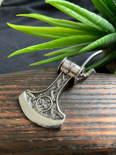 Lying on a wooden surface with a plant in the background is stainless steel silver coloured pendant in the shape of an axe head. With detailed patterns and a black sun rune symbol engraved onto it this item is stunning.