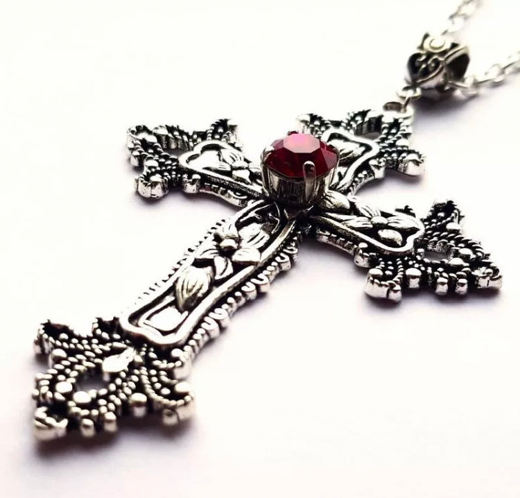 A close up view of a cross pendant which is silver in colour with a red stone embedded into the middle of it. Off of the cross are patterned pieces and on the cross itself there is an intricate flower design.