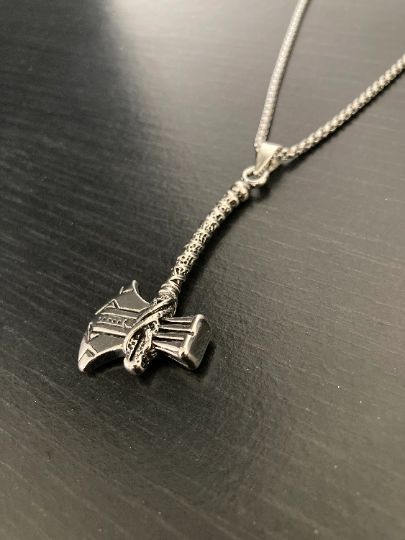 An angled view of a axe pendant on a chain which is shiny to look at and incredibly detailed. It features a long handle and is beautiful as well as solid and weighty. 