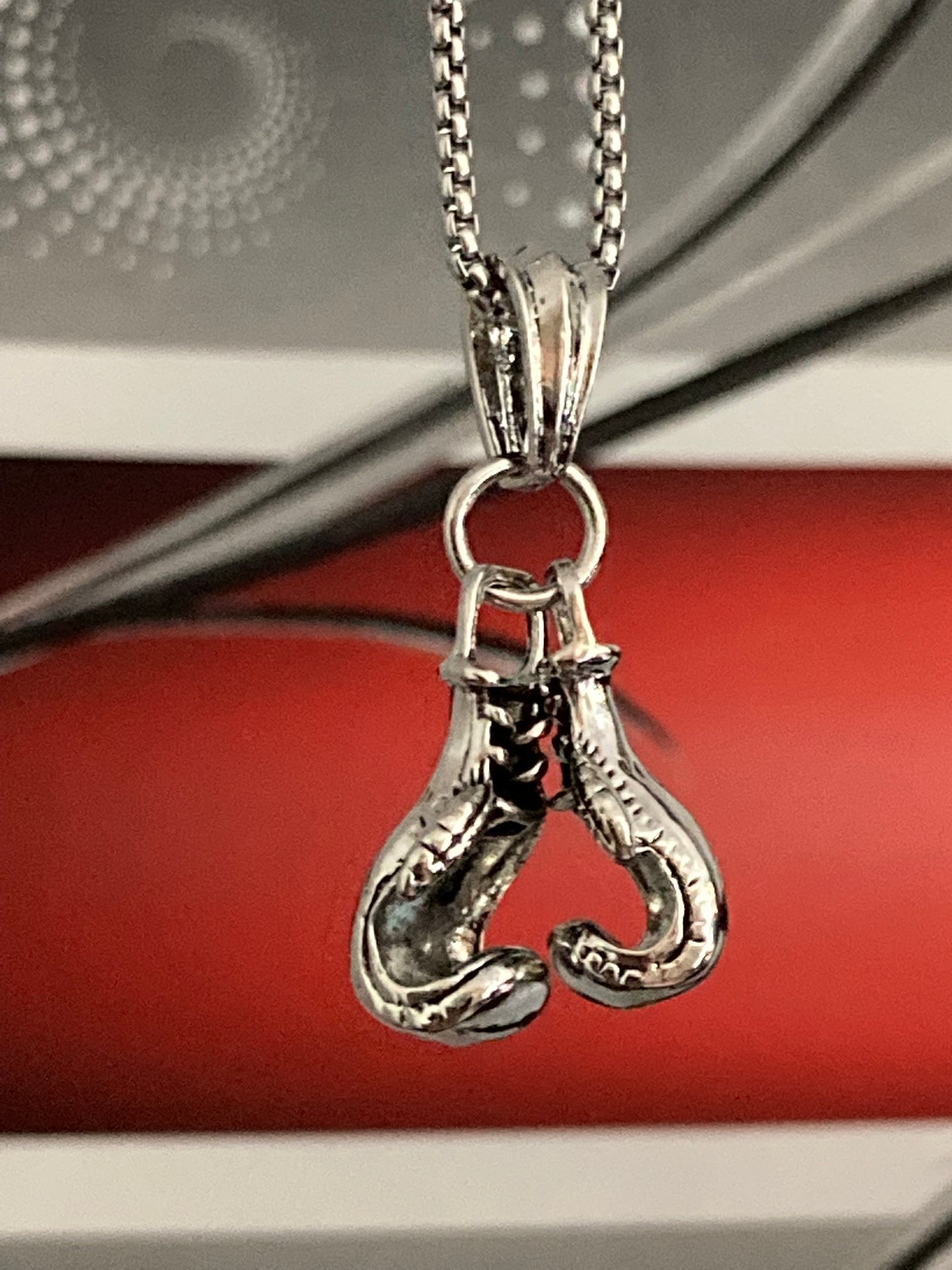 A grey and red background is the host for a silver coloured pendant and chain. This is of a pair of boxing gloves which are small but solid with detailed markings and a ring holding them onto the chain.