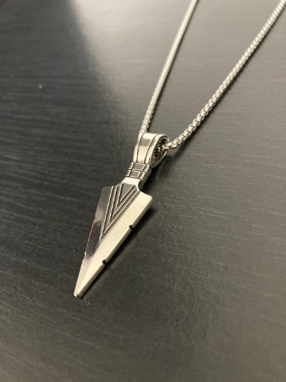 A black grainy surface holds an arrow shaped pendant attached to a chain. Both are the colour silver and are shiny to look at with the arrow having v shaped lines etched onto it.  