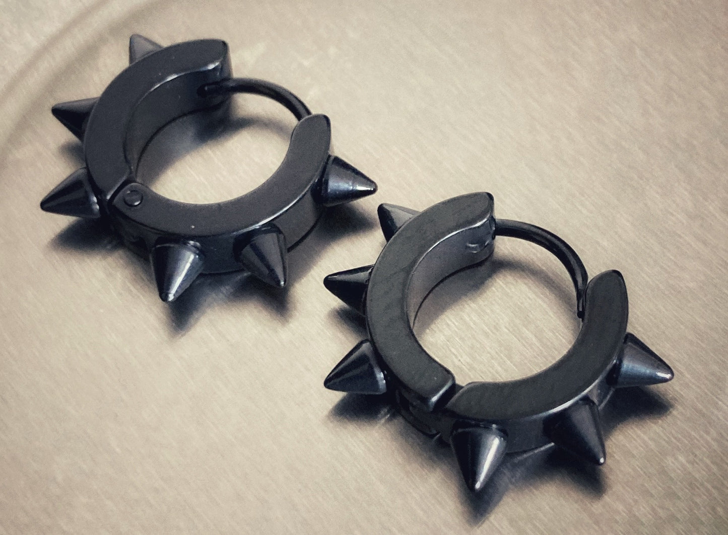 Two black round earrings sit on a grey surface. These are black in colour and shaped like a ring. Spikes are attached to the outer part of them giving them a gothic look. Shiny to look at and lightweight to hold.