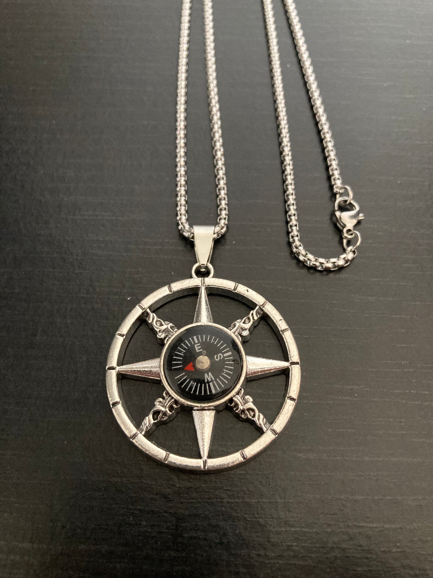 A view from above of a fully working compass necklace. Attached to a stainless steel chain the pendant is a circle shape with an outer ring which has eight cut out points leading from there to the centre where the compass sits.