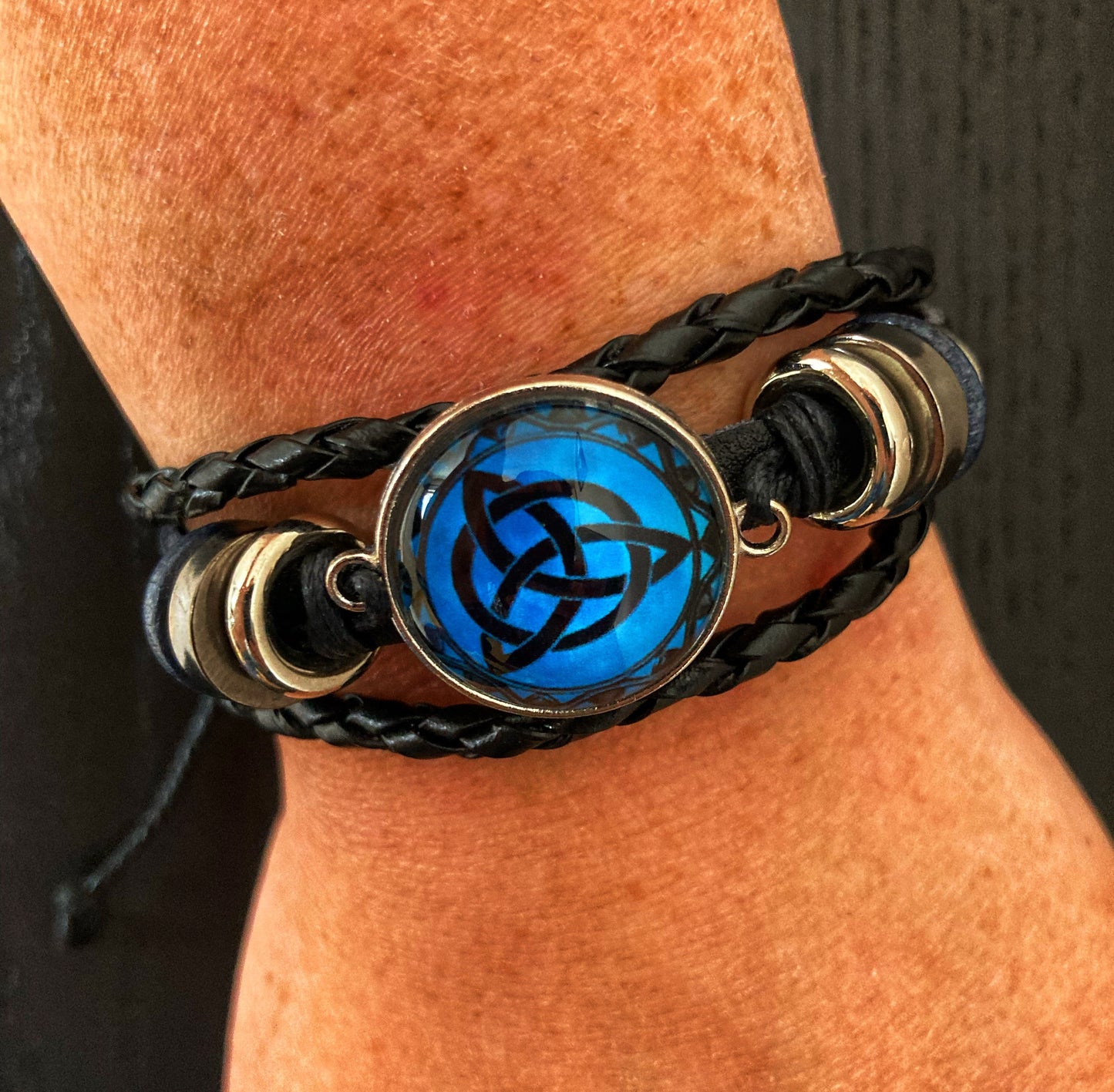 A close up of a freckled wrist wearing a leather bracelet. Black in colour are the 3 bands that make this up with a central feature that has a metal round ring and then a bright blue background with a triqueta design in black printed on it. This is covered by a clear glass looking cover.