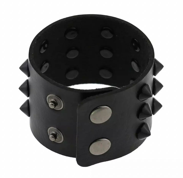 On a white background sits a black wide bracelet which is covered in spikes all around it. At the forefront you can see the press studs for fastening which look to be solid and sturdy.