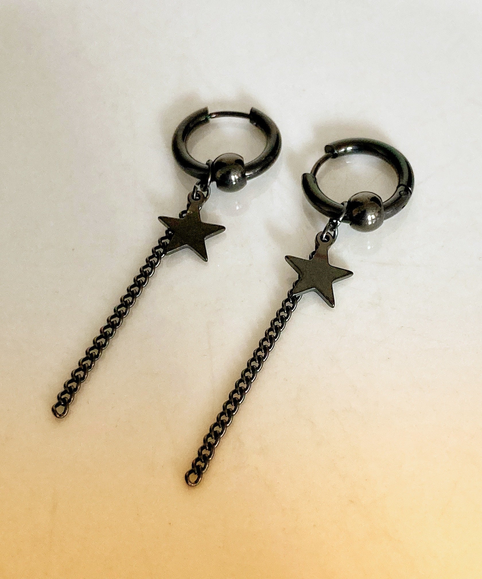  A pair of earrings lie on a white surface. They are black in colour and are a thin ring shape with a linked chain hanging from them and also a star. They are stainless steel and feel like a quality item.