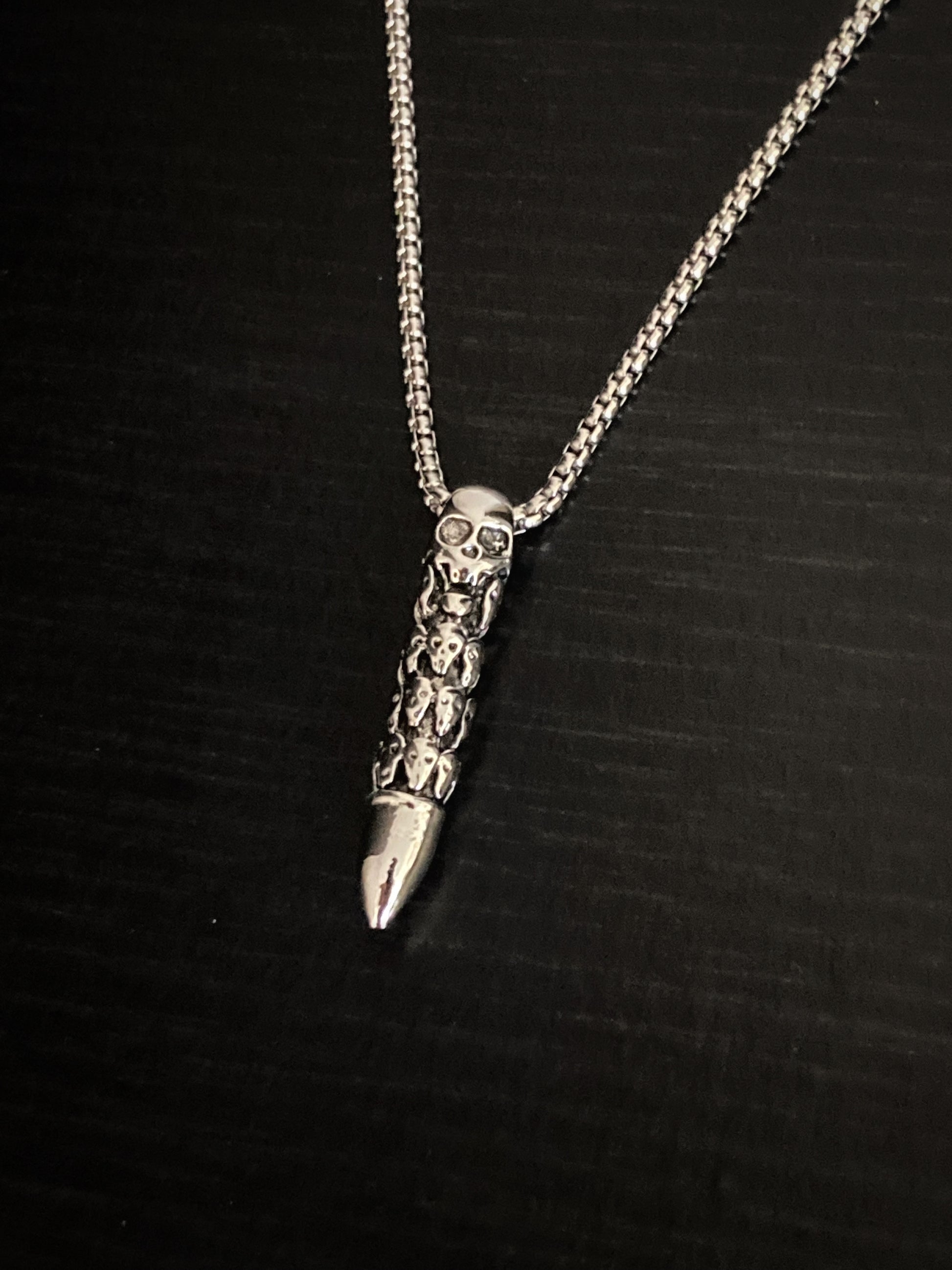 A view from above of a skull carved bullet pendant on a chained necklace both of which are silver in colour and made of stainless steel. The skull heads are intricately engraved onto the main body with the tip being left plain.