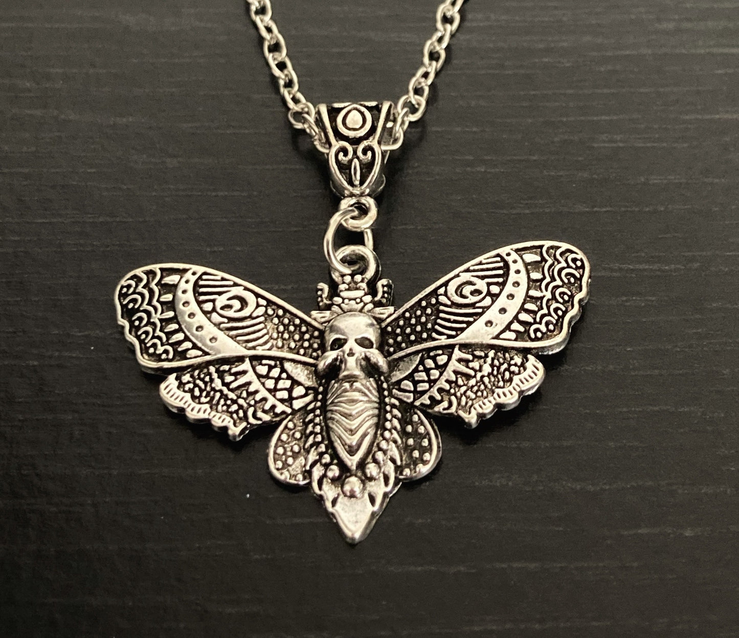 A close up of a death moth shaped pendant silver in colour. The main body of the moth features a skull design and the wings are a beautifully detailed design that cover the surface of them. When holding this item you can feel the quality of it.