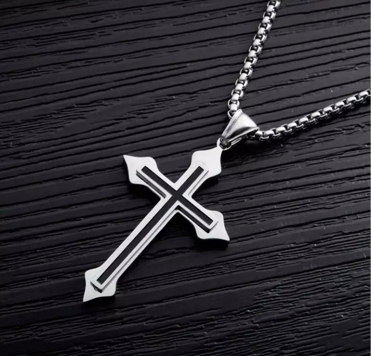 Lying on a black surface is a cross necklace. With a sturdy looking chain the cross has a smaller one on top of it. The chain and larger cross are a shiny silver and the smaller one is black.