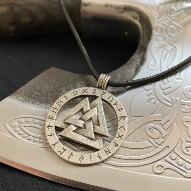 A circular pendant with a valknut design is sitting on a steel axe head. There is a black cord as the chain and the main design is 3 interlocking triangles in a circle with rune symbols round the outside. 