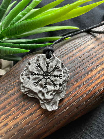 Lying atop a wooden axe handle with green plant in background is a rock shaped heavy pendant wit ha black engraving of a helm of awe viking symbol. The item is silver in colour and is shiny with a black cord necklace attached to it.