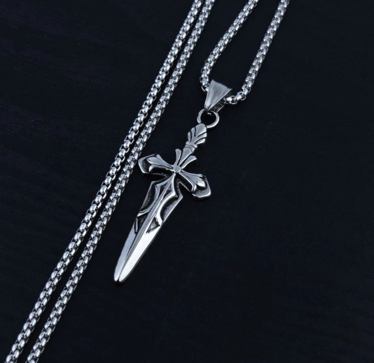 A sword pendant on a chain lies angled on a black surface. There is a cross design to the sword and intricate markings on the cross. The whole thing is a silver colour and it is lightweight to hold and wear.