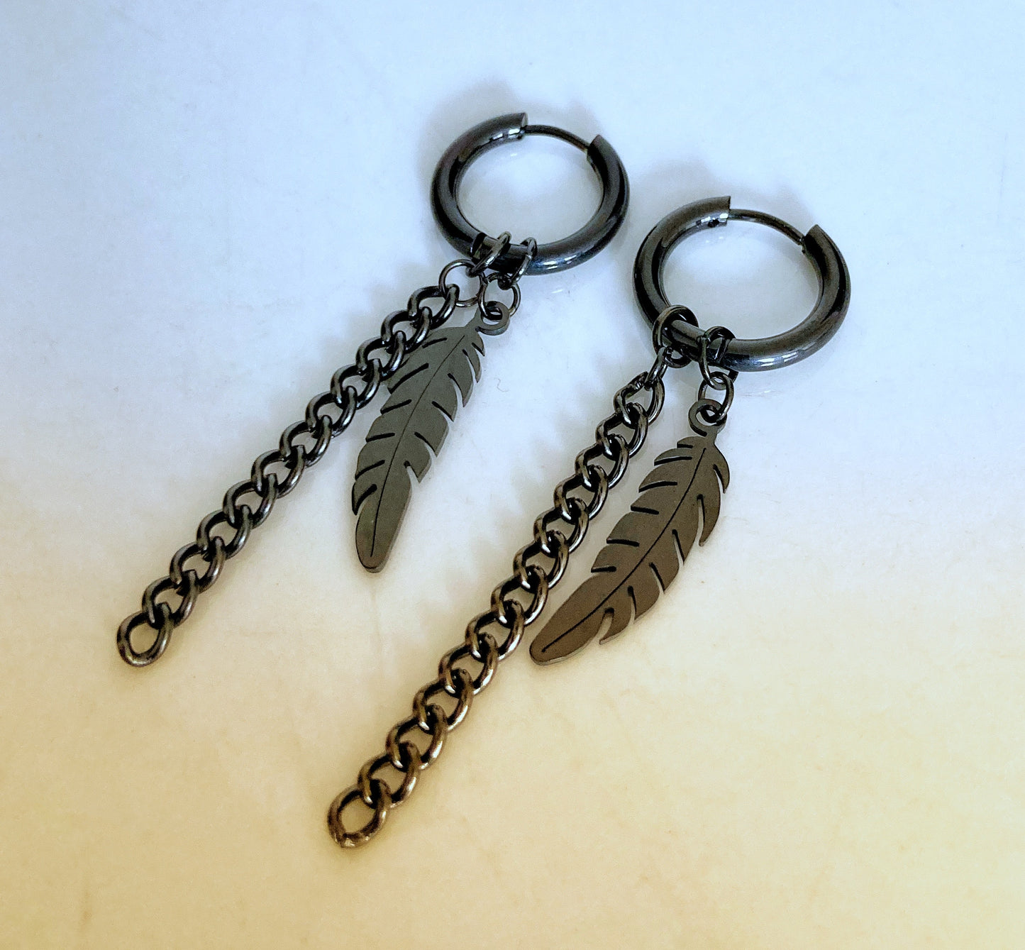 A pair of earrings are lying on a white surface. These are hoops with a feather design attached and next to it a link chain that dangles down. They are very lightweight and made of stainless steel.