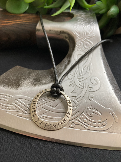 Laying on a patterned axe blade is a stainless steel pendant which is a circle inscribed with black runic symbols around the outside. Tied with a knot is a black corded necklace holding the item in place.  