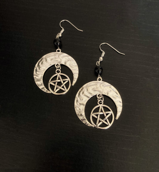A pair of earrings lying side by side. These are silver in colour and are the shape of crescent moons with a pentagram hanging from the inside curve of the moon. 