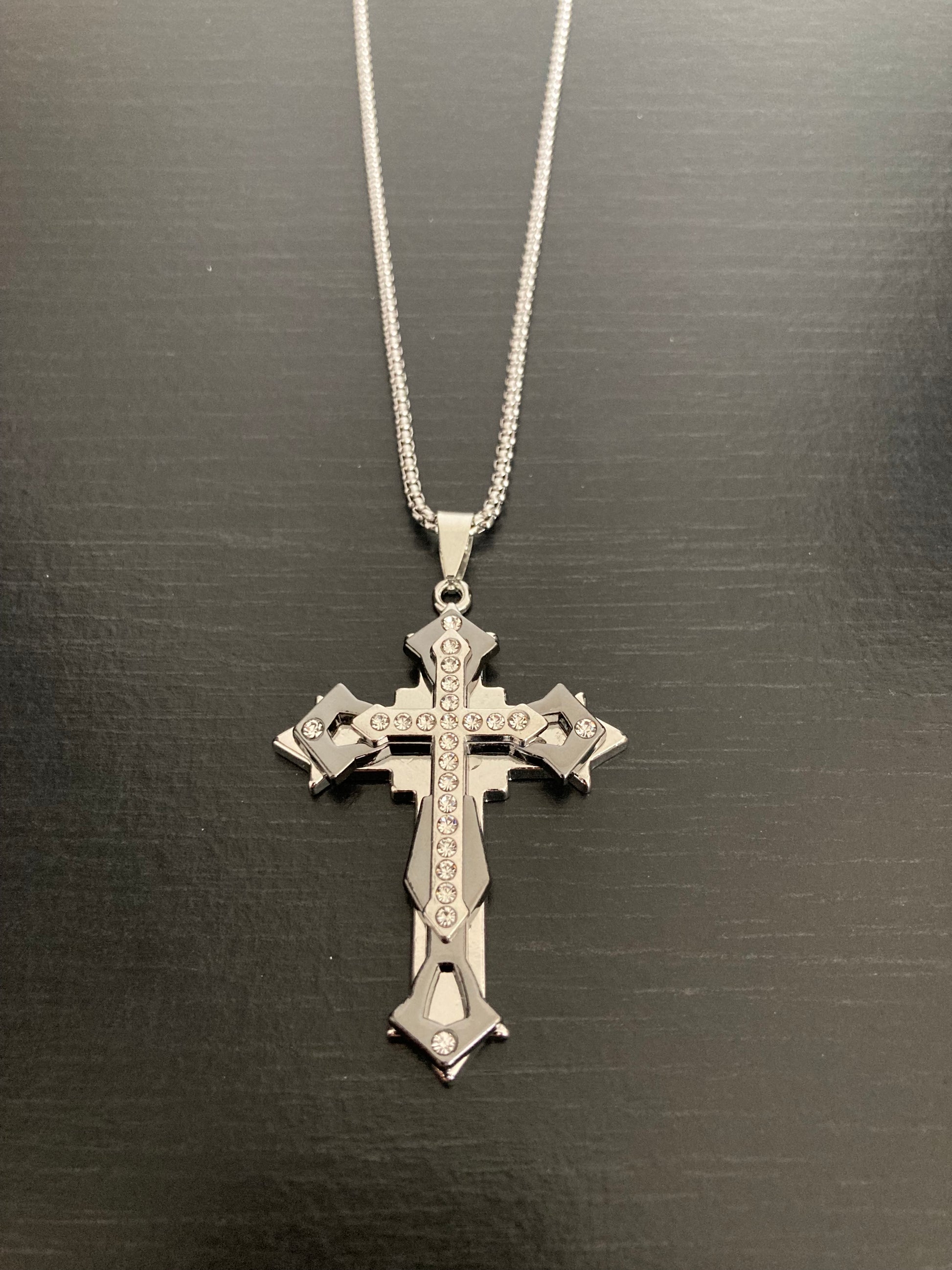 A necklace is on a black surface that is silver in colour. There is a sturdy looking chain with a pendant attached that is made up of three crosses on top of each other. It is slightly gothic in design and chunky looking with a shiny appearance.