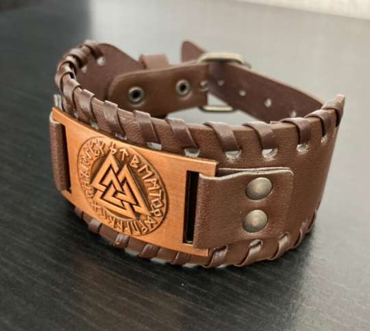 An angled view of a brown leather cuff bracelet on a black background. The bracelet is fastened showing how it would look once worn. There is a design of valknut and runes engraved onto a bronze metal plate which is detailed and of high quality.