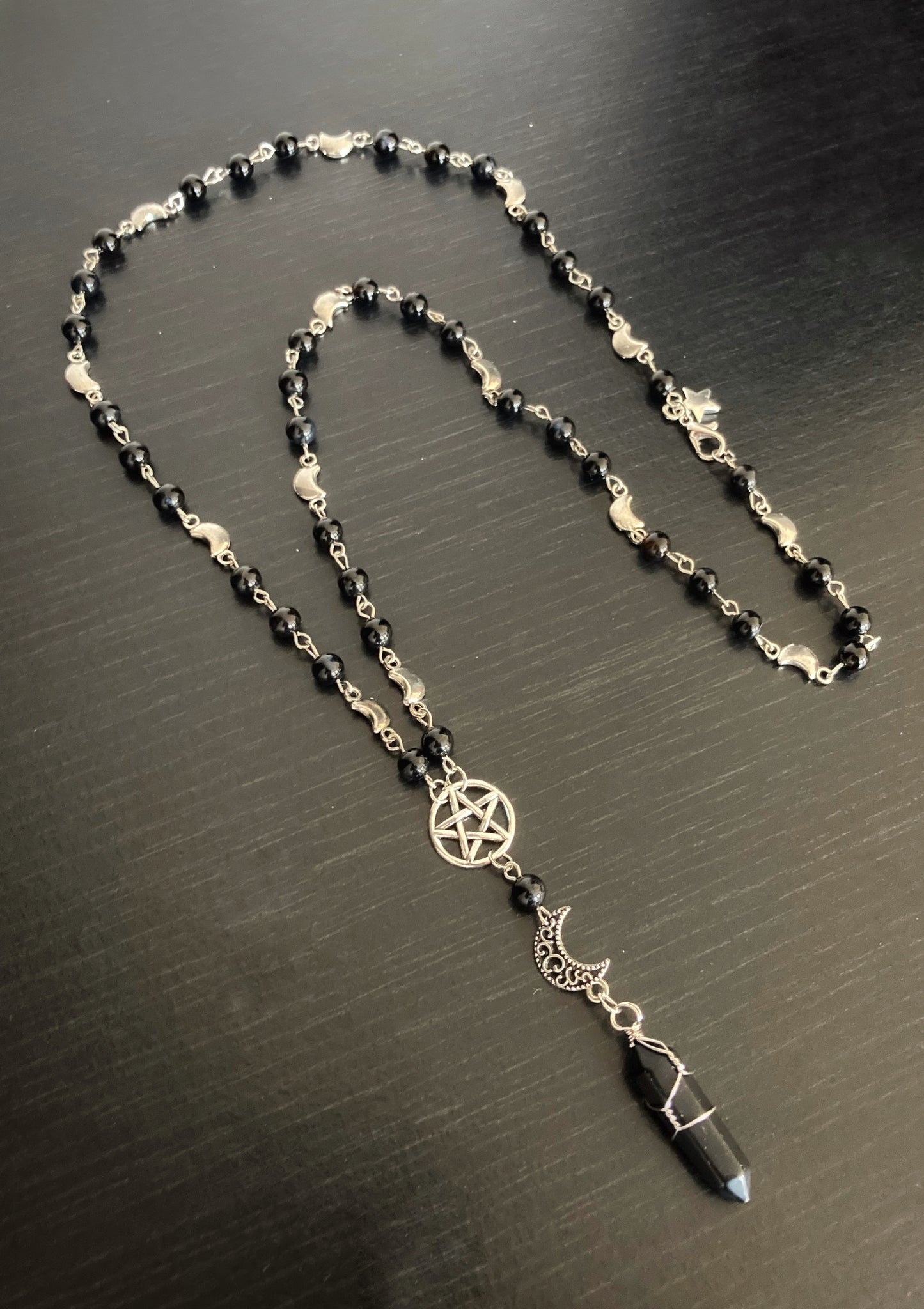 A black onyx stone hangs down with a silver coloured crescent moon and a pentagram design above it. The necklace itself is a mixture of small black shiny beads and a silver coloured chain with a metal clasp for fastening.