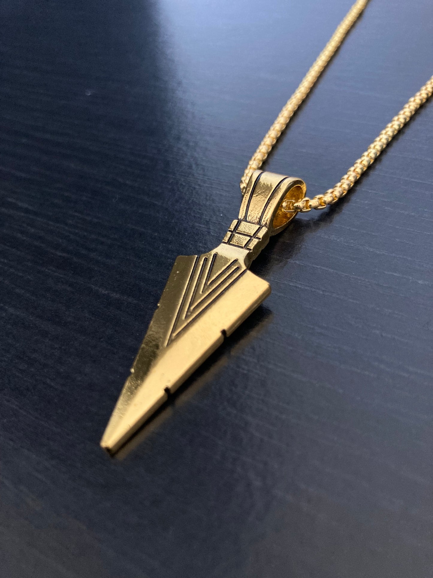 A gold coloured arrow head pendant attached to a chain of the same colur lies on a black surface. There are engraved v shaped markings on it and it is made from stainless steel. It feels light in weight to hold and wear.