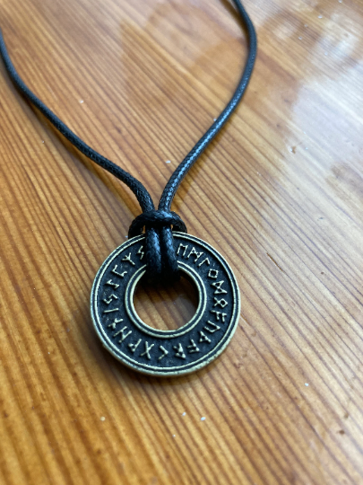 On a brown wooden table sits a black cord necklace that has a round disc shape attached to it. This has a hole in the centre of it and there are markings of runes all the way around it.