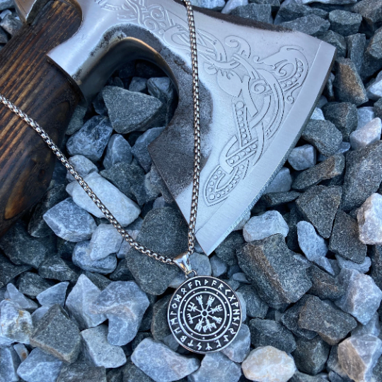 Necklace is laying on small grey stones and is draped across a wooden axe with a steel blade. The pendant is round and black with a silver colour compass pattern in the middle and runes round the edge which you can feel if you run your hand over it.