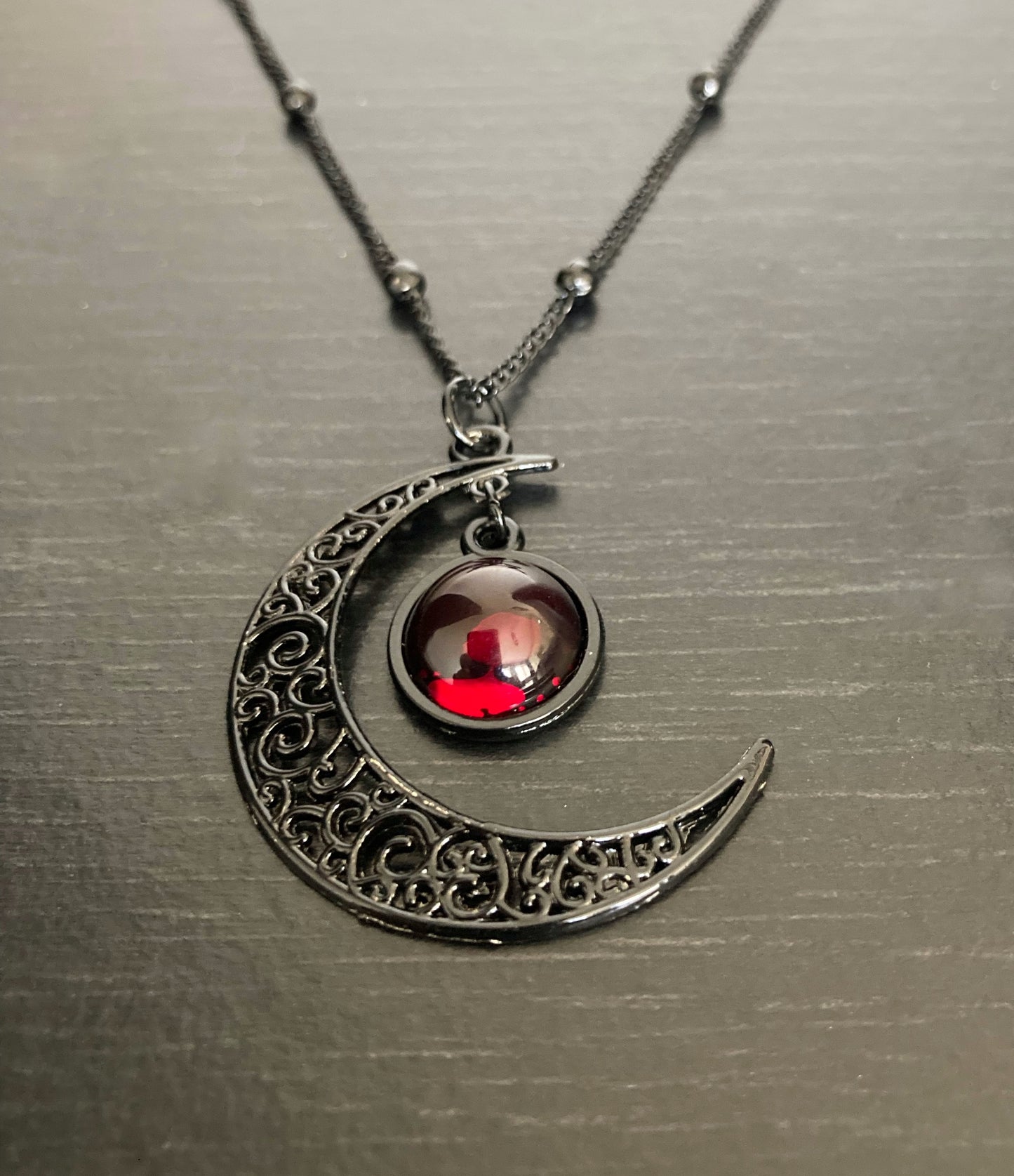 A red glass circle is hanging into the curve of a crescent moon necklace. The chain is thin with solid small ball shapes dotted along it. The moon and chain are black in colour and a lightweight stainless steel.