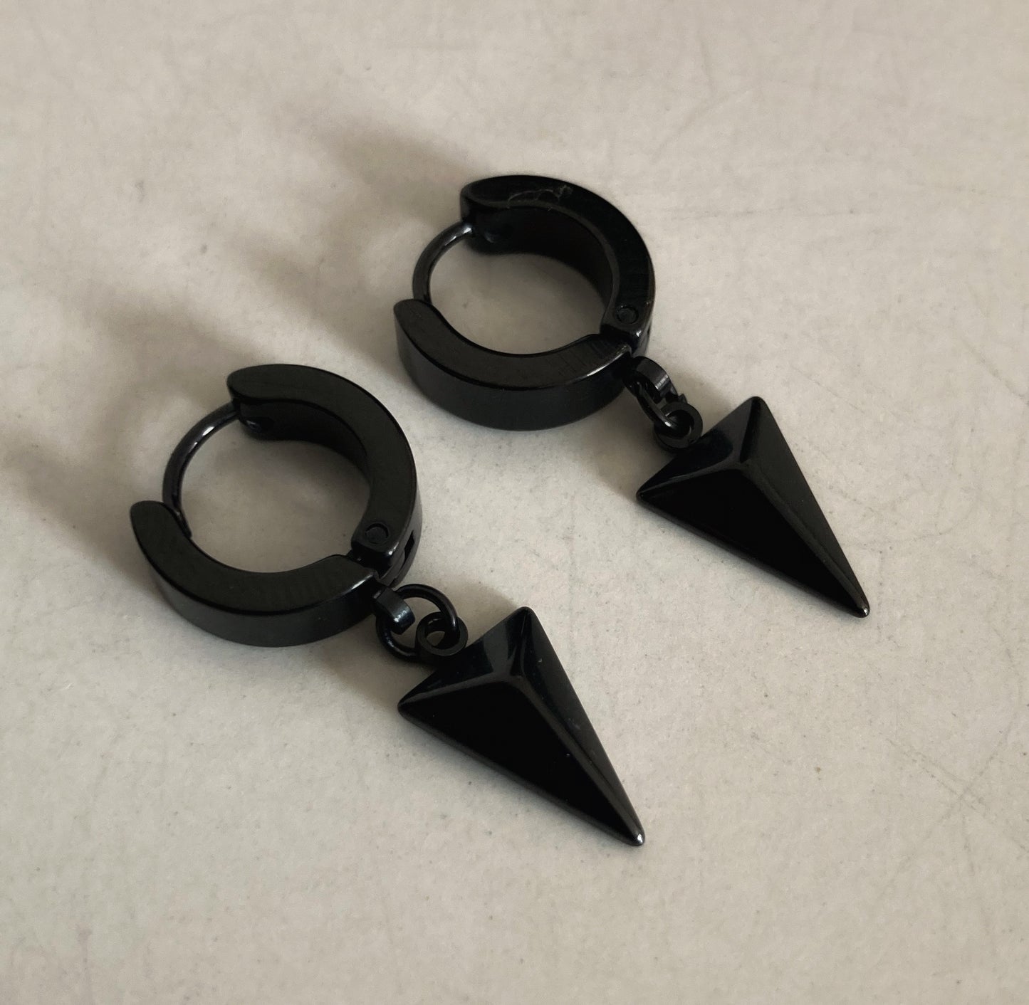 A pair of black earrings lying side by side. They feature a 3D triangle shape dangling from a chain attached to a ring shape earring that can be opened to go in your ear.