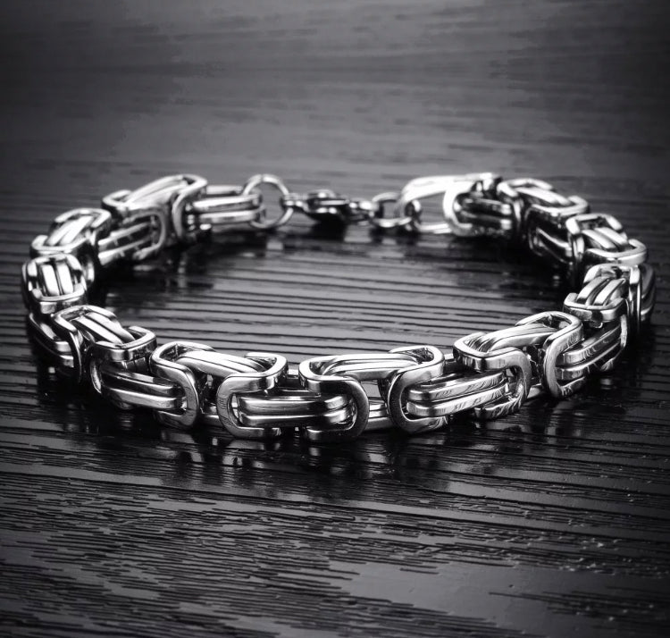 Upon a textured black background sits a silver coloured bracelet. This is made up of interlocking chains and is chunky but light in weight. There is a metal clasp that fastens the item and it is shiny in appearance.