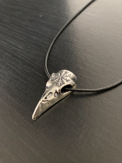 A raven bird skull necklace is lying on a black background. It features a black cord with metal clasp and is made of stainless steel. It is a hollow pendant and is therefore one sided in design. The cord runs through the hollow eyes of the skull.