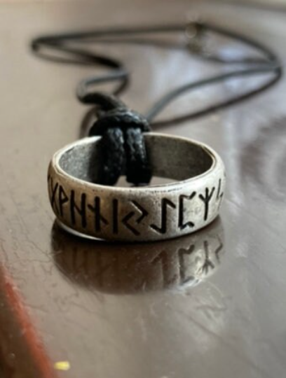 A close up view of a rustic looking silver coloured ring necklace sitting on a brown wooden surface. There are rune letters inscribed around the outside of the ring which are black. The ring is attached to a black cord which looks sturdy. 