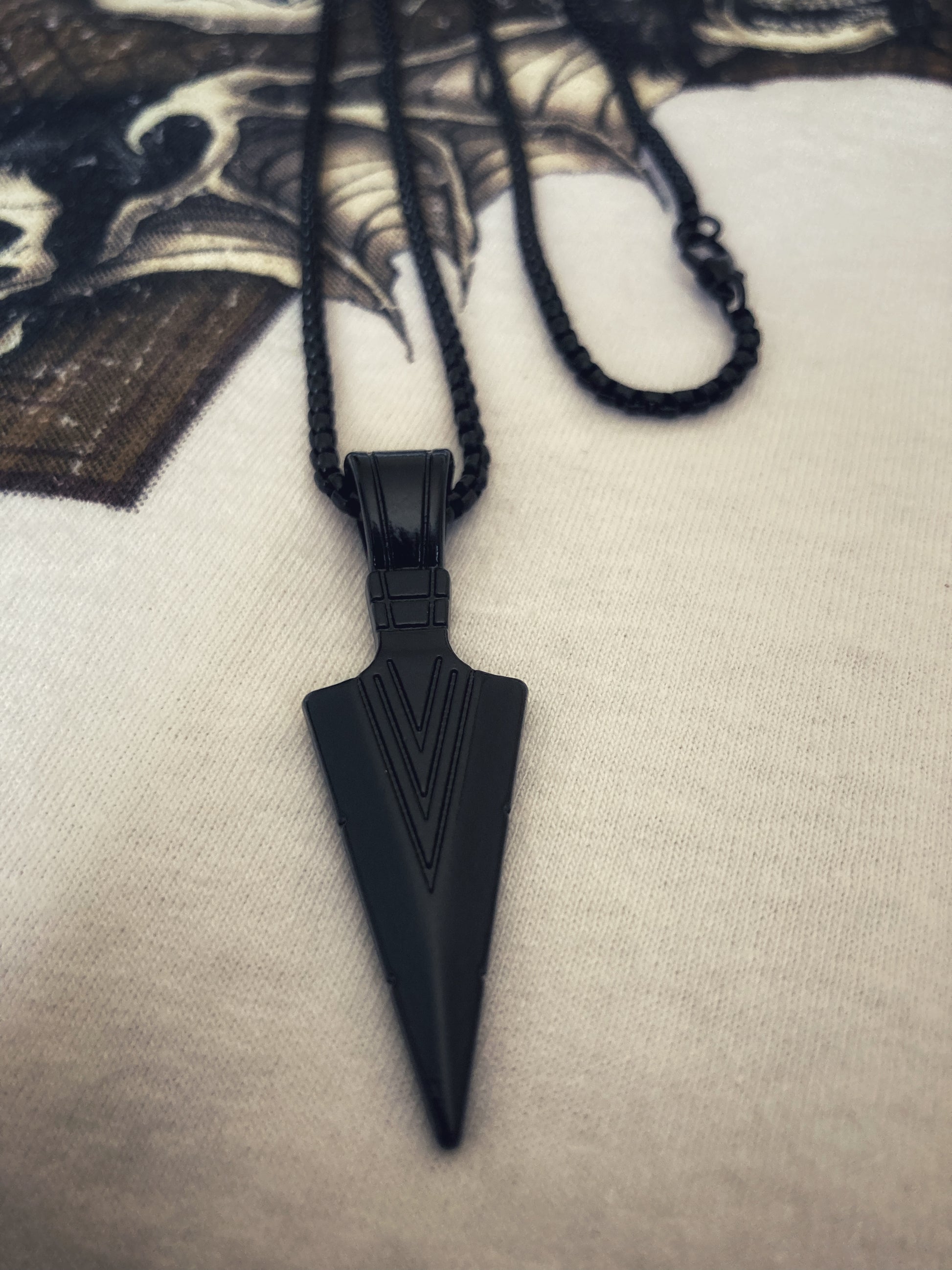 A black arrow pendant is shown here with the design being of the head of an arrow. A chain is attached and both items are black stainless steel. When you touch it you can feel the lines that are engraved as a pattern on the item. 