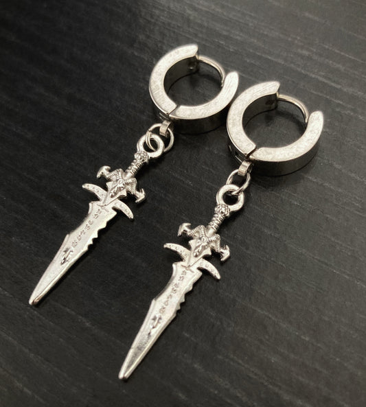 A pair of sleeper earrings sit on a black surface. They have small daggers attached to them that dangle down and are silver in colour.