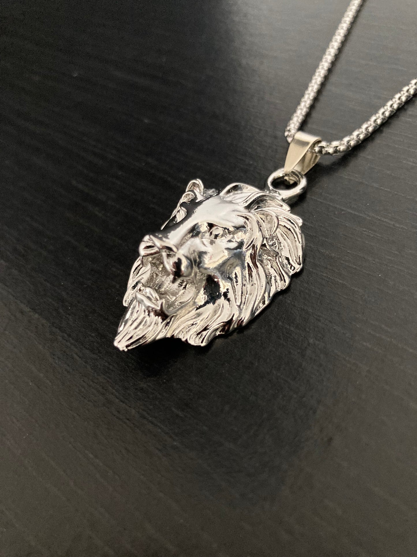 On a black background lies a detailed lion head pendant attached to a sturdy looking chain. Both are silver in colour and made of stainless steel with a shiny appearance. The face of the lion is detailed and the item is chunky and light to hold.