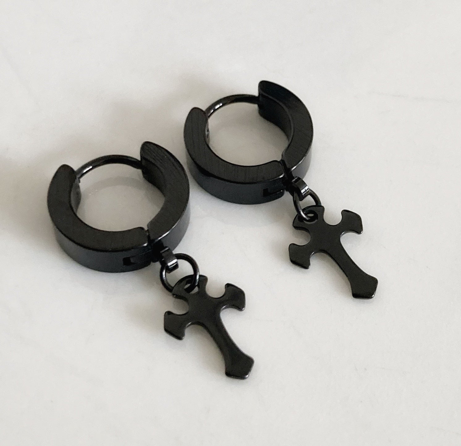 A pair of earrings lying next to each other. They are both black in colour, round in shape with a black cross attached to them. Made of stainless steel these glisten in the light as they have a shiny appearance.