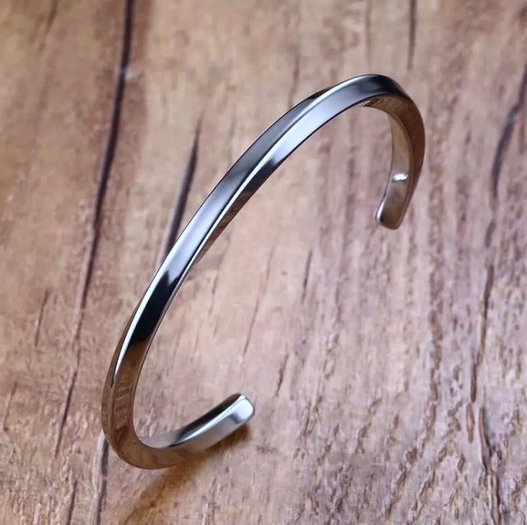 On a wooden surface sits a stainless steel bracelet that is thin and silver in colour. This is more of a bangle style and is twisted in its design. There is an open section for sliding onto your wrist and the item is very shiny in appearance.