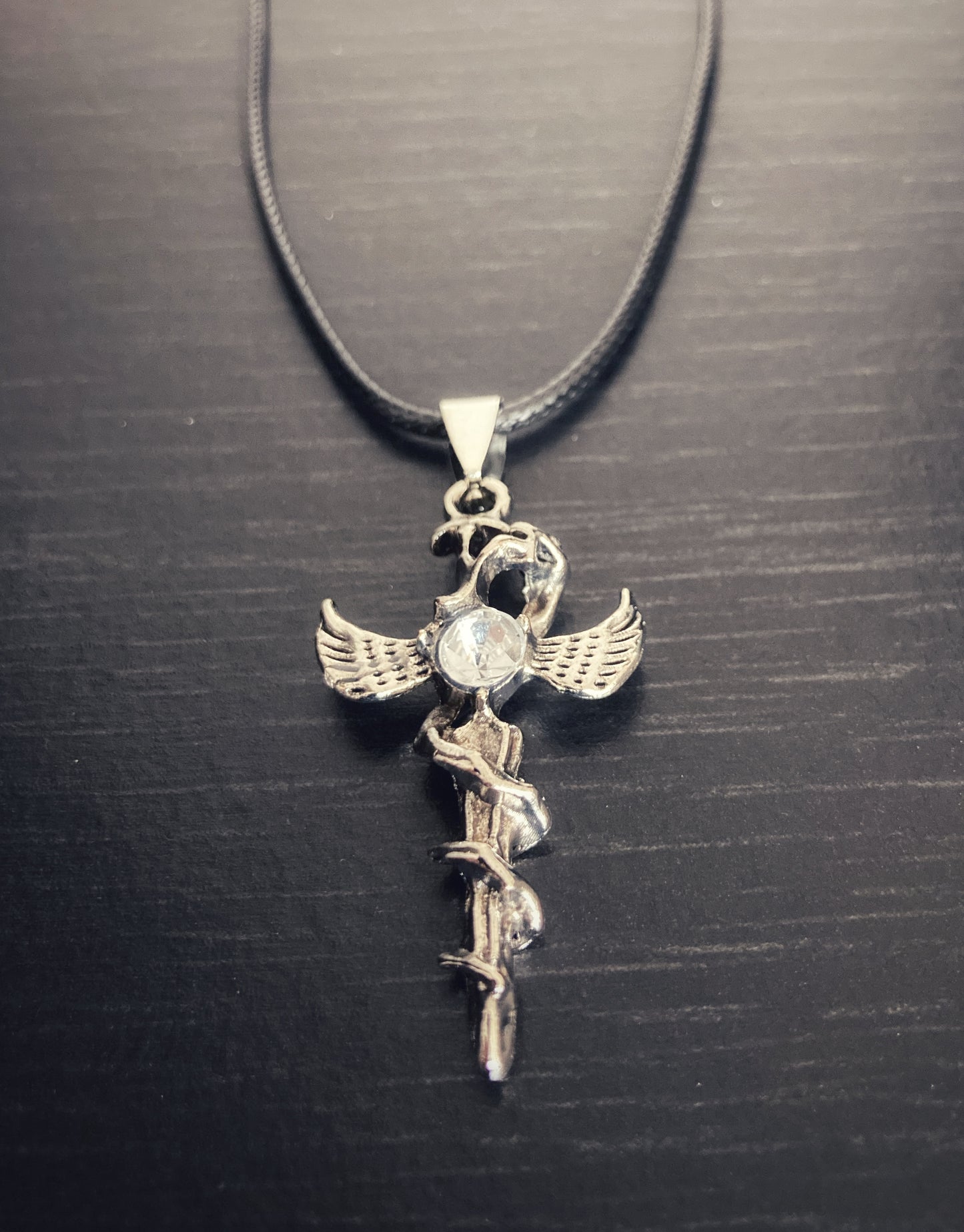 On a black background sits a corded necklace that is black with a dagger shaped pendant which is silver coloured. There is a clear glass stone embedded onto the hilt of the item and there is a serpant wrapped around the length of the dagger. 