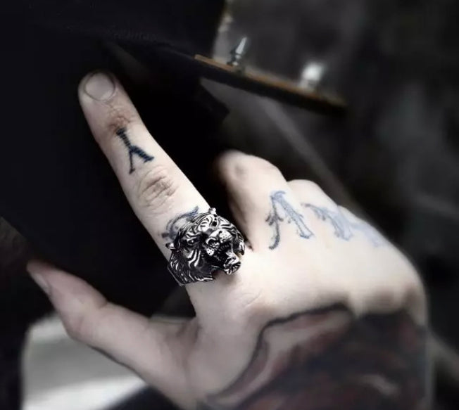 A tattooed finger is wearing a chunky looking tiger head ring which is silver in colour. This has detailed markings on the face of the tiger and feels solid when wearing it.