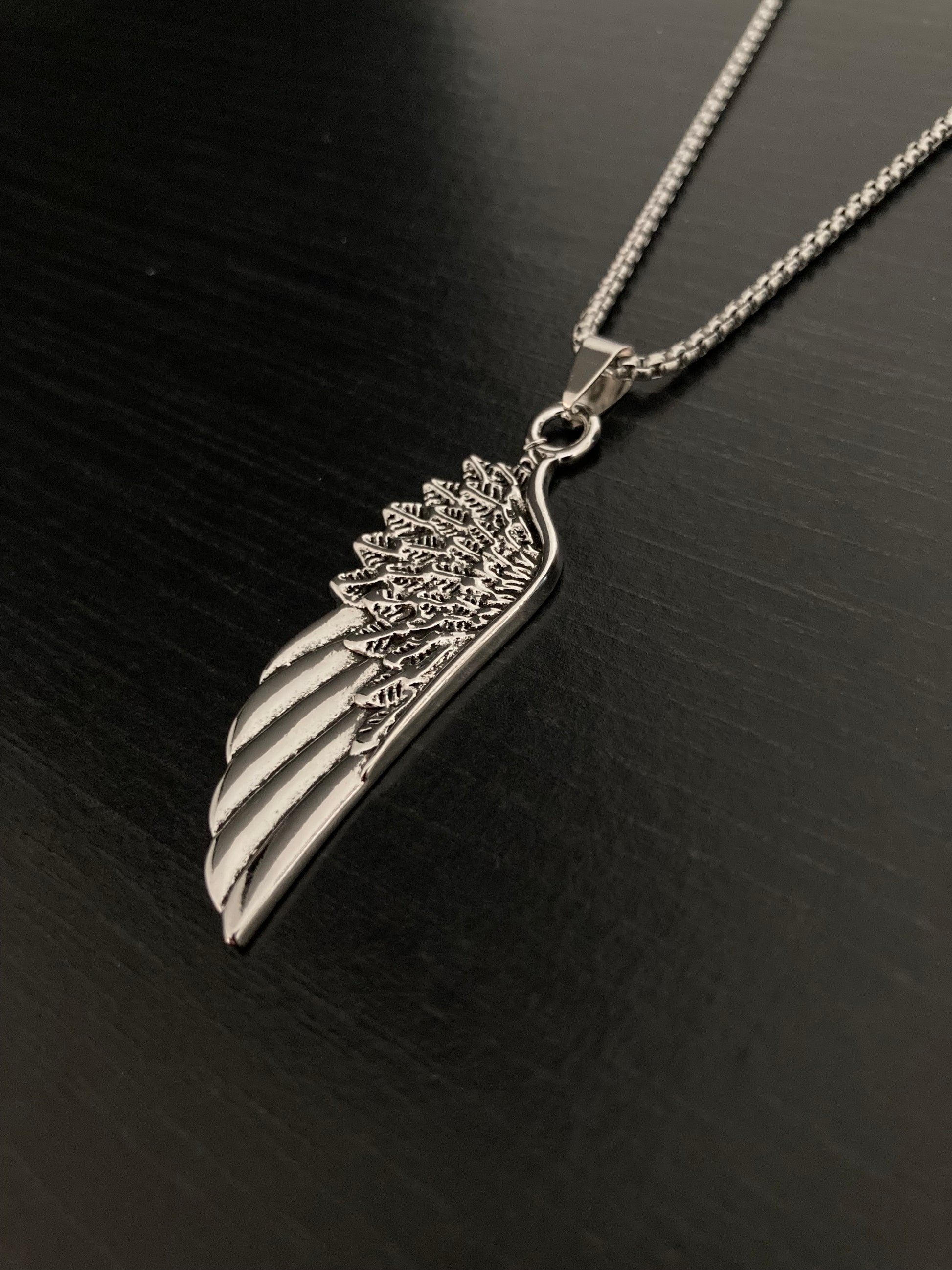 A silver coloured wing pendant sits on a black surface. With detailed feather like markings and made of stainless steel this looks stunning. It is attached to a chain thats feels like a quality item and sturdy. 