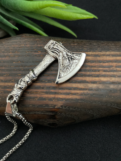 A close up of a pendant in the style of a viking axe is lying on a wooden item. This stainless steel piece is in the shape of a axe and when you hold it, it feels solid. It is shiny to look at and had a patterned hadle as well as a design on the head