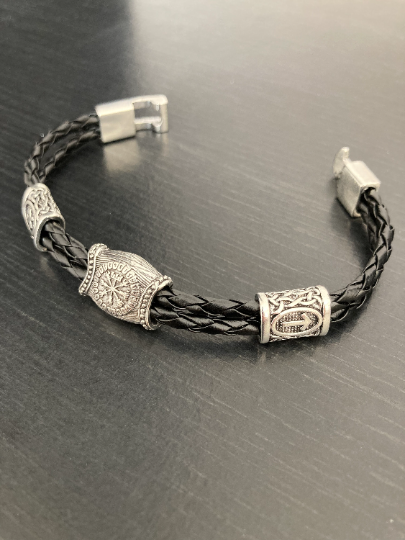 A bracelet featuring silver coloured rune pieces is on display. The main rune has a vegvisir engraving and these are attached to a black leather band. There is also a solid looking fastening piece.