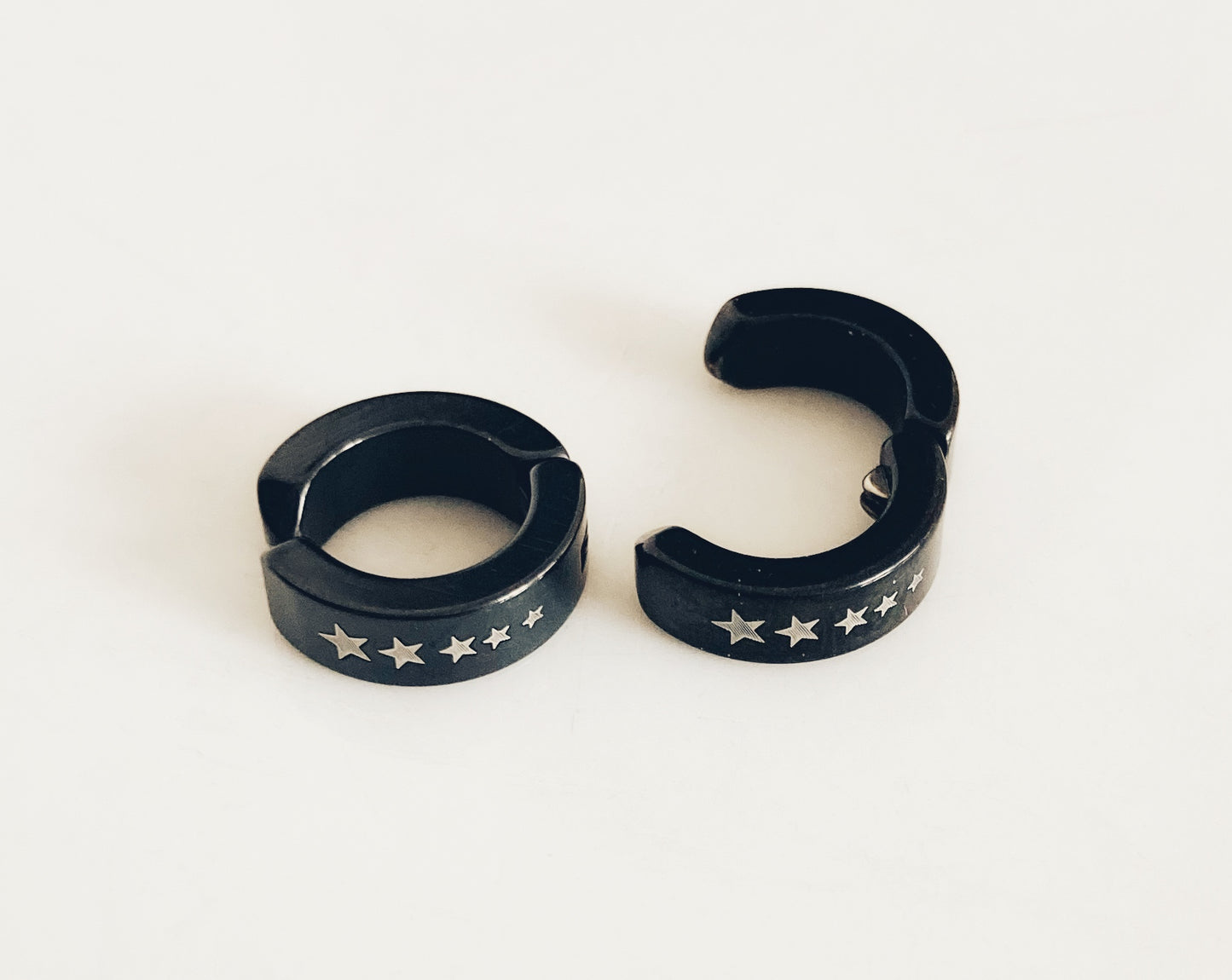 Two earrings sit next to each other. Both are black with white stars and one is opened to show how wide it will go to clip onto an ear. The other is more closed showing the ring style shape of it.