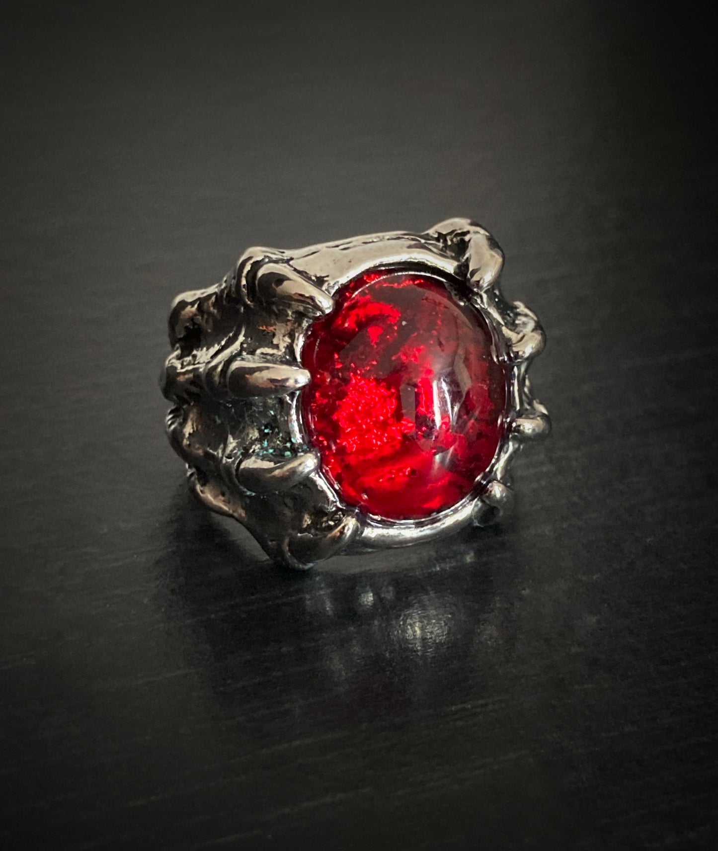 A silver coloured ring sits on a grainy black surface. This is made to look like a dragon claw that is holding onto a red stone which is made of glass. This is a chunky item that feel weighty when on your finger.