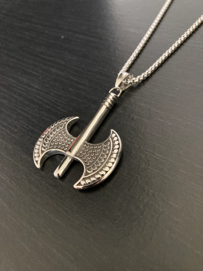 A slanted view on a black surface of a necklace which is silver in colour and features an axe pendant. This is a battle axe style which has two identical heads ataached to a handle and is weighty and solid in the hand.