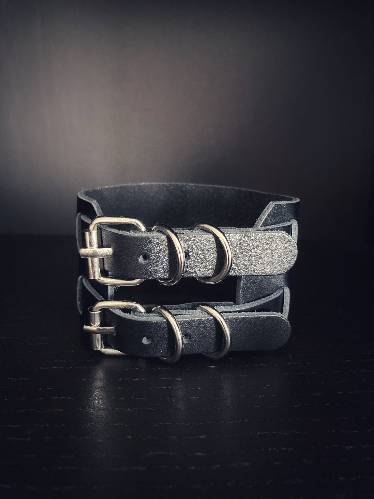 On a black surface sits a leather bracelet that is wide and black in colour. You can clearly see the two metal buckles that fasten this with several holes for making it different sizes.