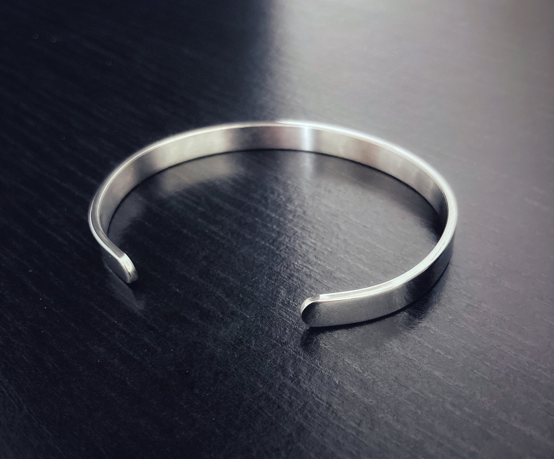 Sitting on a grainy black surface is a shiny silver coloured plain bracelet. It is fairly narrow in width and there is a gap for sliding onto a wrist making it adjustable.