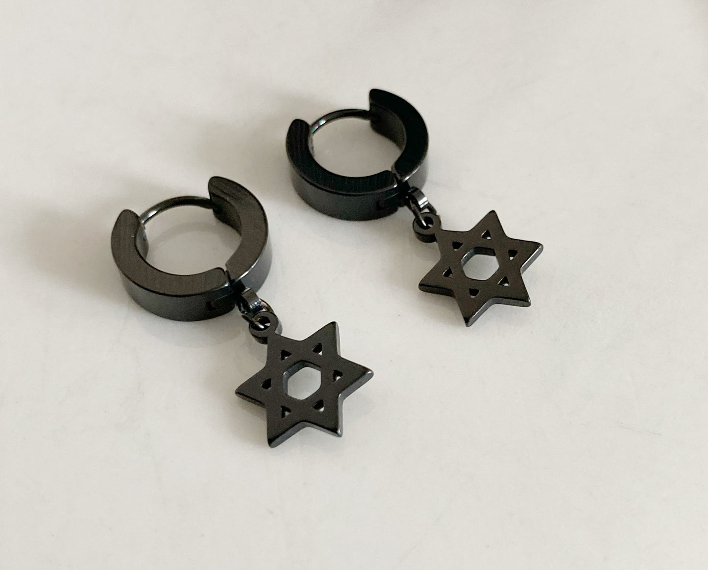 A pair of star of david shapes hang from round earrings. They are black in colour and made of stainless steel. They have a slight shiny look to them and are lightweight to hold.