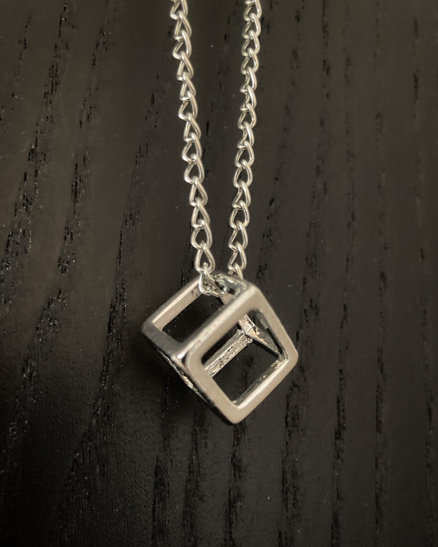 A shiny silver coloured pendant in the shape of a three dimensional cube is leaning on a black background. The hollow cube is attached to a chain and both are stainless steel.