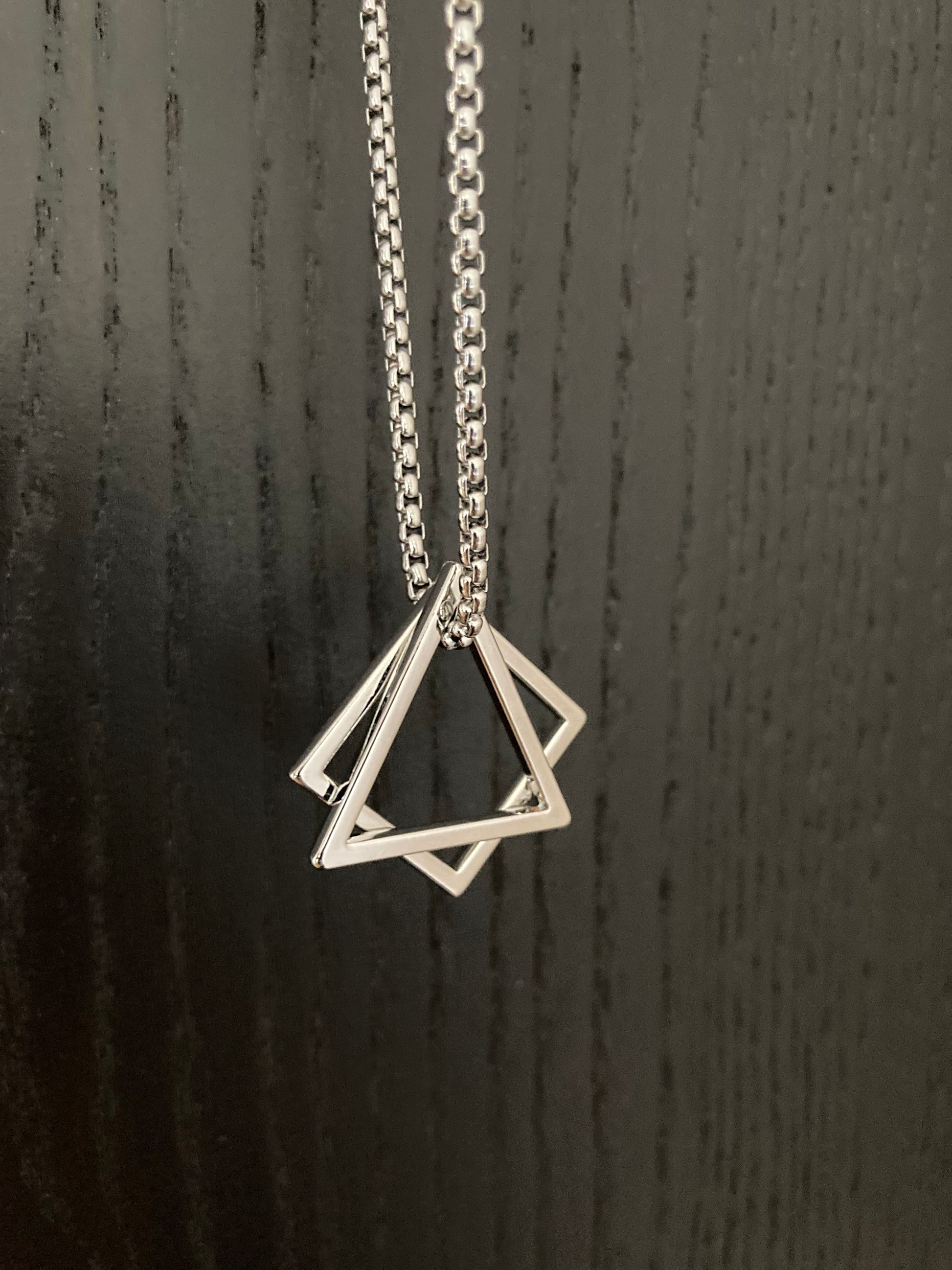 Against a grainy black background is a silver coloured necklace hanging up. Attached to the chain is a hollow shaped triangle and a hollow square. These are shiny in appearance and very light in weight. The chain is sturdy and of high quality.