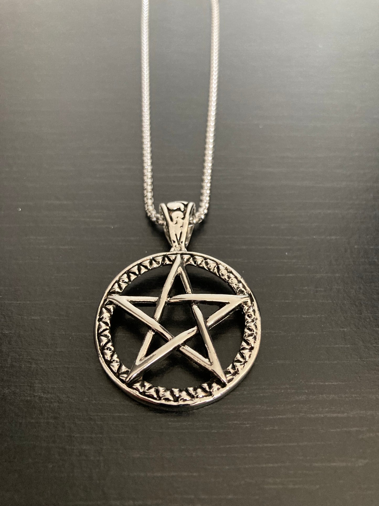 A silver colour circular pendant is sitting on a black background. Inside the outer circle is a pentagram cut out shape. A sturdy looking chain is attached to the pendant and both are made from stainless steel.